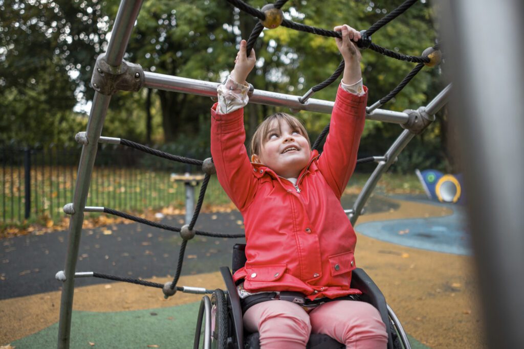 A low-angle close-up of a little girl who is also a wheelchair user having fun in a playground in a public park in Newcastle upon Tyne. She is having fun as she pulls herself along with the rope frame.